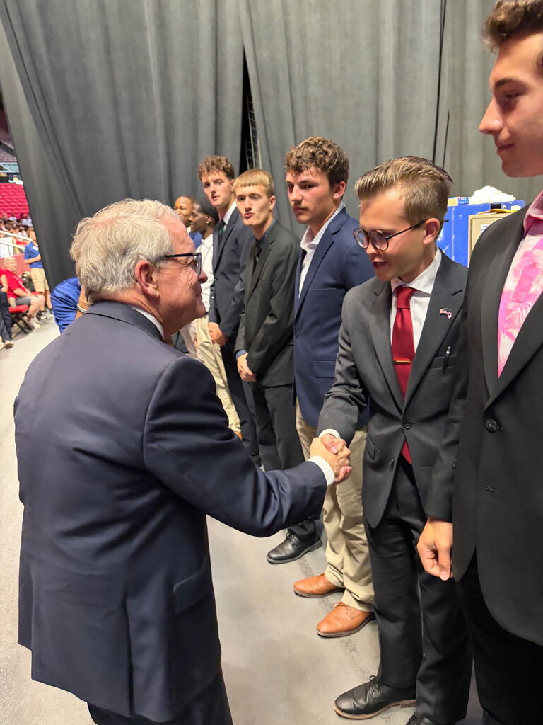 Several young men stand in suits and ties; one older man shakes hands with a man in front of him.