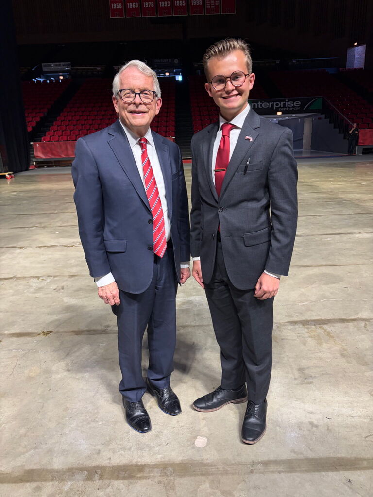 Two men in suits and ties, one with glasses, stand in a vast, empty arena with red seating.