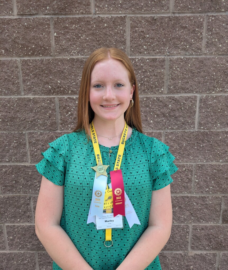 Woman wearing a green polka dot blouse and three ribbons, smiling for a photo against a brick wall.