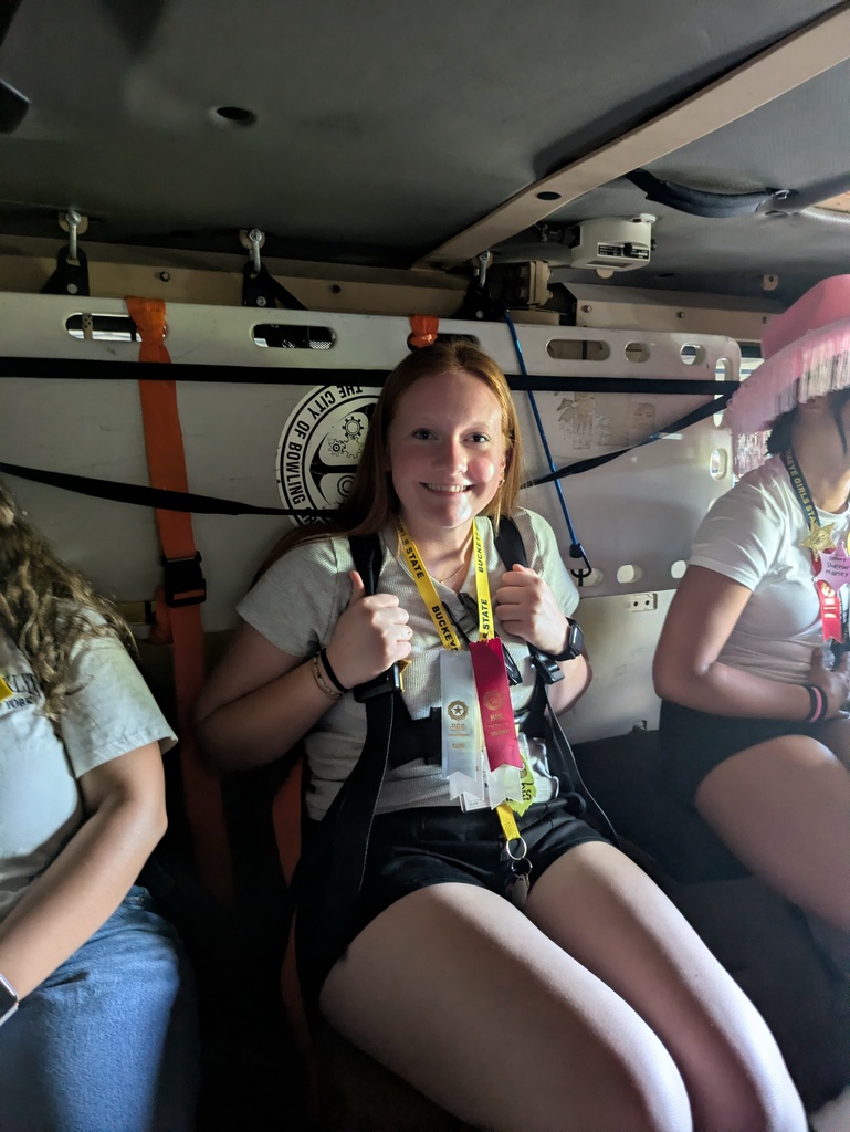 Three women seated inside a vehicle. The woman in the center wears a harness and holds a badge.