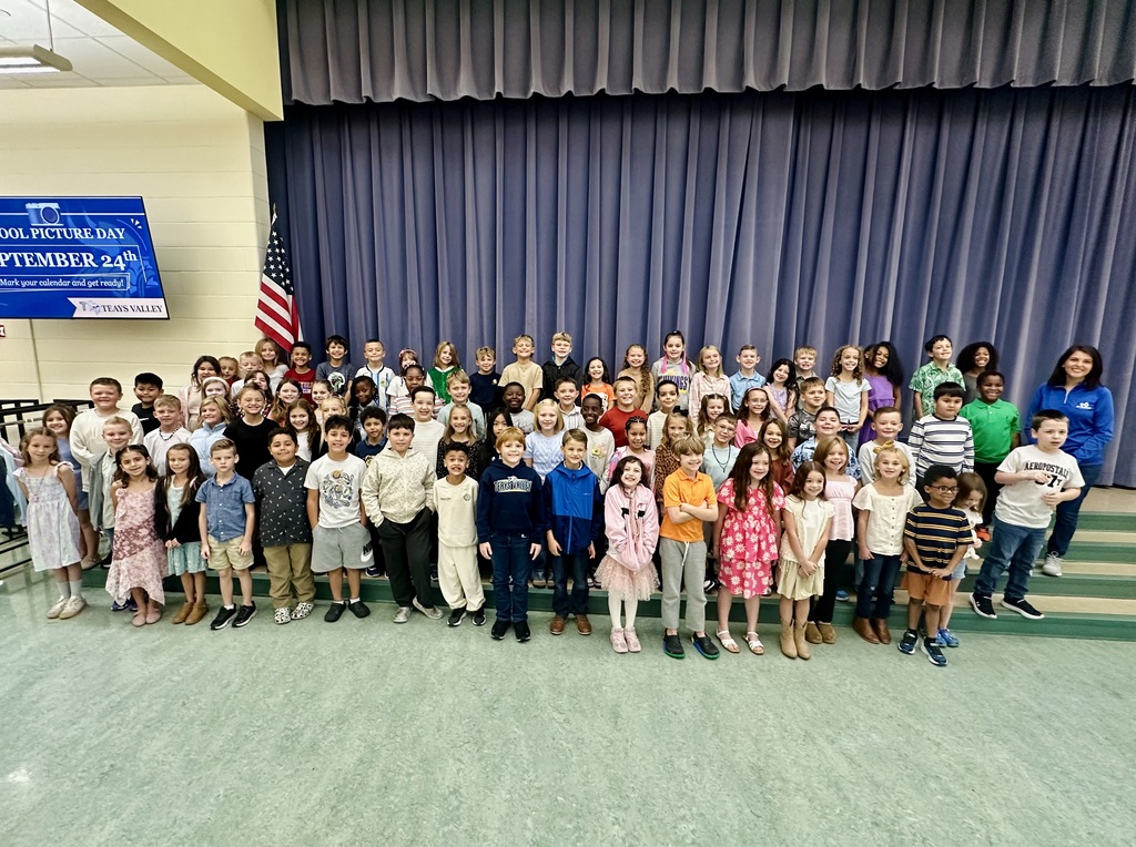 Many children stand in rows in a room, possibly at a school, with a blue banner and flag.