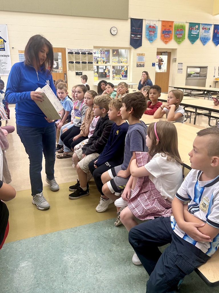 A woman reads a book to a group of children sitting at a long table in a school cafeteria.