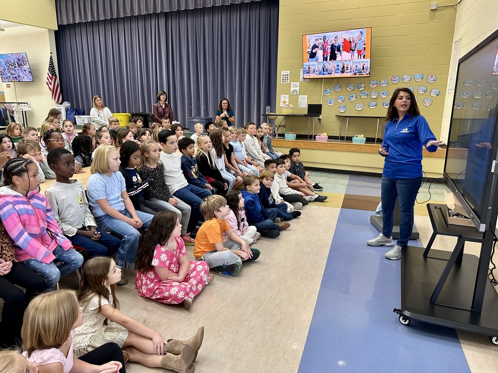A woman in a blue shirt presents to a group of children sitting on the floor.