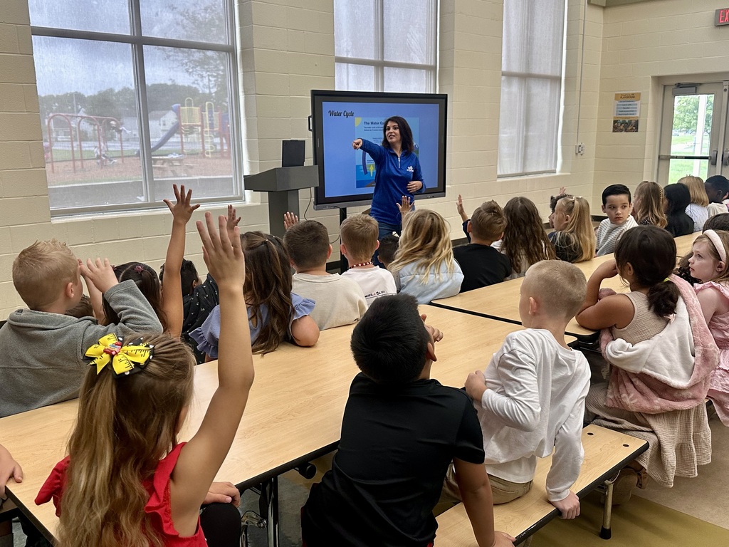 Classroom with children seated at tables, a teacher at the front using a large monitor.