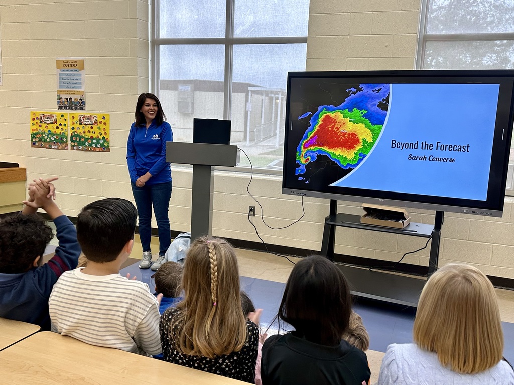 Woman in blue shirt at a podium with a screen, teaching students about weather. Students seated.