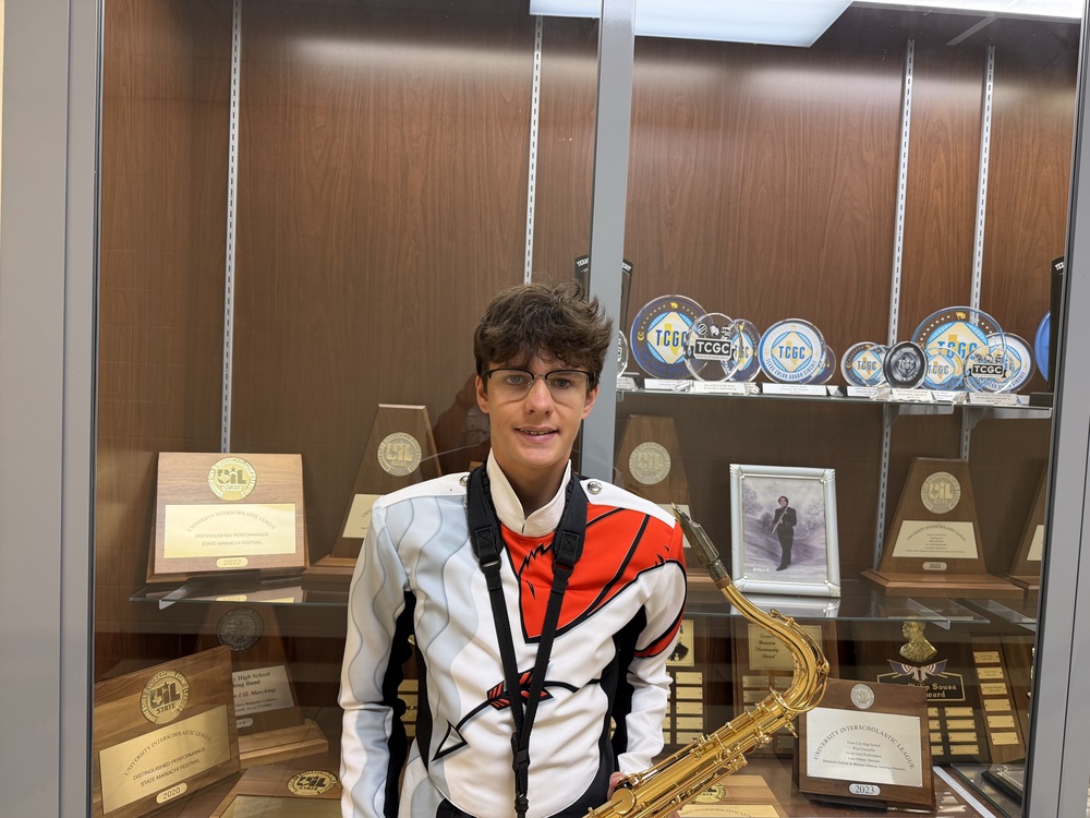 Band student in uniform poses with instrument in front of case of band awards