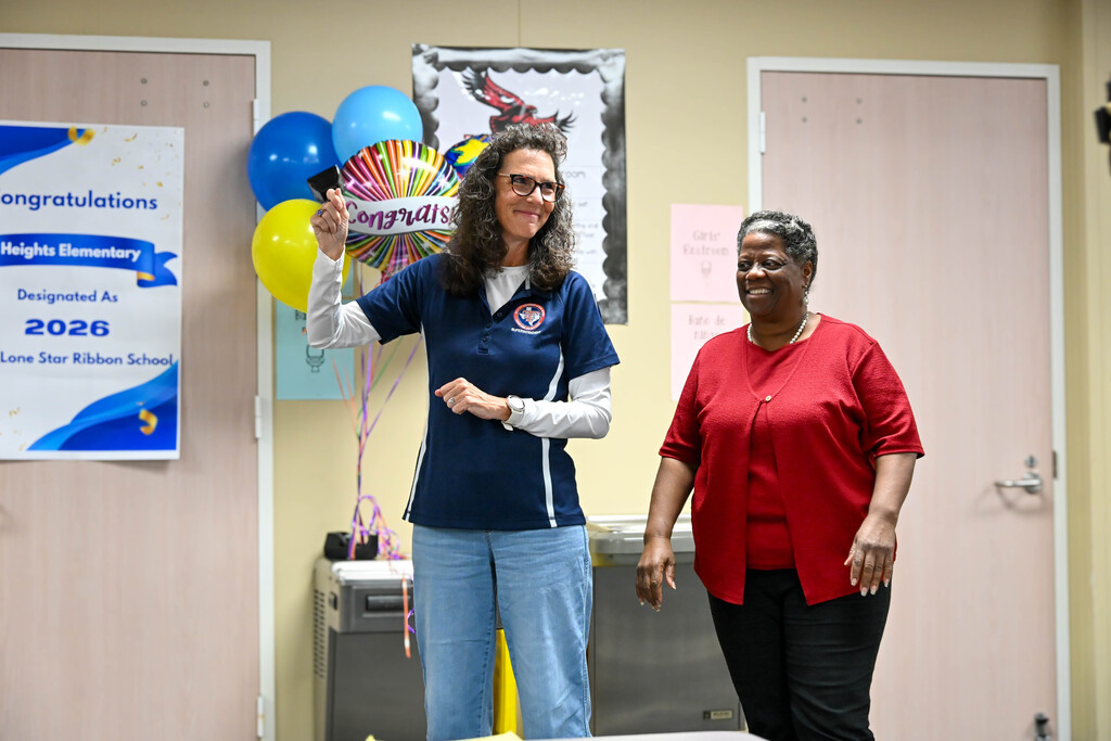 Two education leaders smile during the announcement that Heights Elementary was designated a 2026 Lone Star Ribbon School. A congratulatory sign and balloons are displayed behind them.