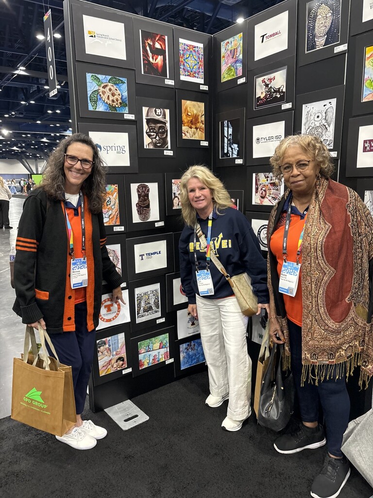 Superintendent and two board members stand in front of a wall of art while superintendent points to the one by LMHS artist.