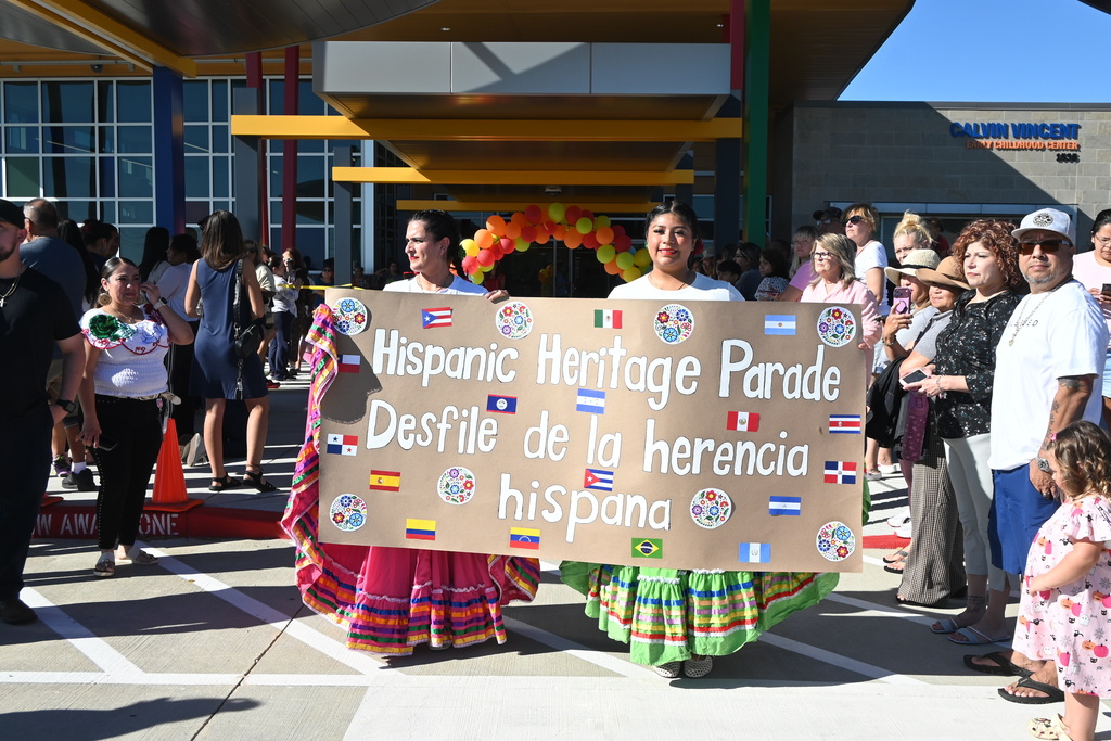 Two employees hold a banner stating "Hispanic Heritage Parade Desfile de la erencia hispana" with painted flags from various countries