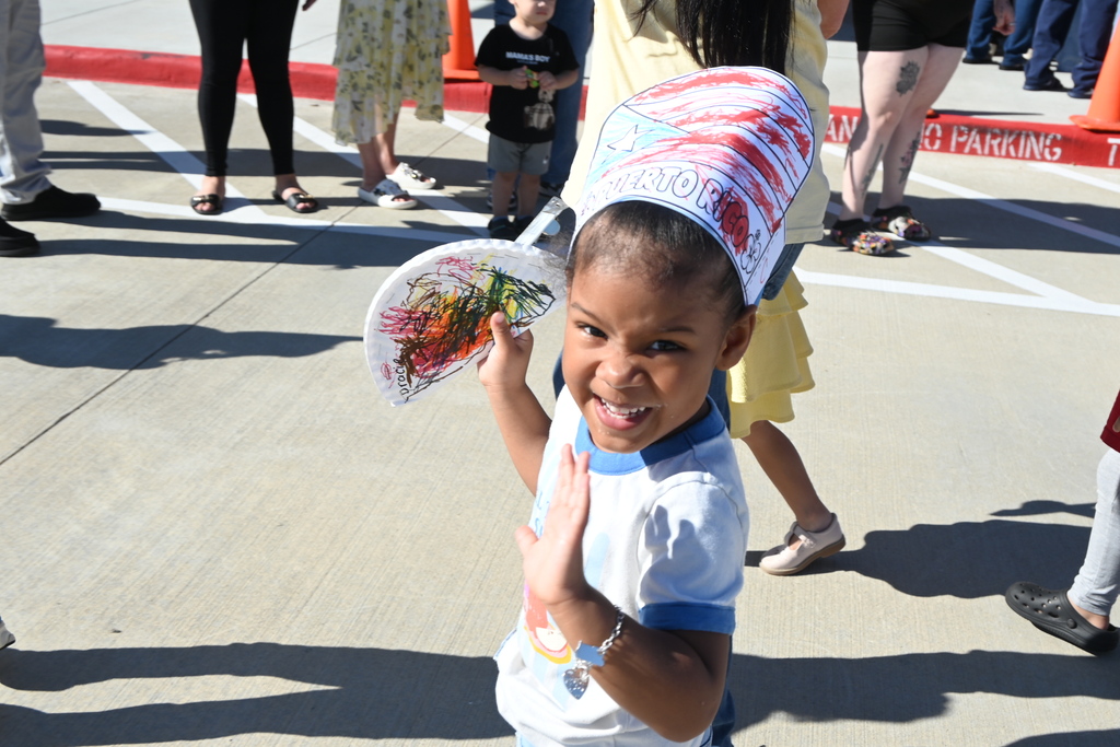 A student holding a paper plate decorated and folded in half and used as a noisemaker and wearing a handmade hat with the Puerto Rican flag colored by the student