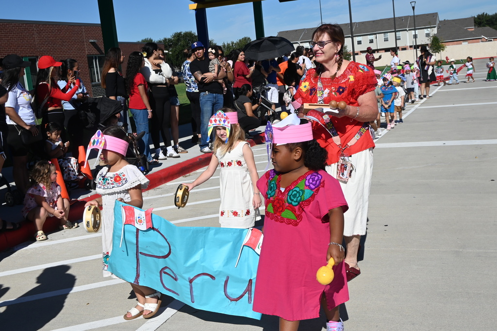 Students wear colorful dresses with flowers while holding a blue banner that says Peru while also wearing handmade paper hats that look like traditional Peru headgear. The students colored their own hats.