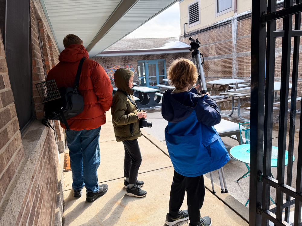 Three people stand under a covered walkway looking out into a school courtyard. Two children in winter jackets hold cameras, while one adjusts a tripod, appearing to prepare for taking photos. An adult in a red jacket stands beside them. The courtyard has picnic tables, patches of snow, and a brick building in the background.