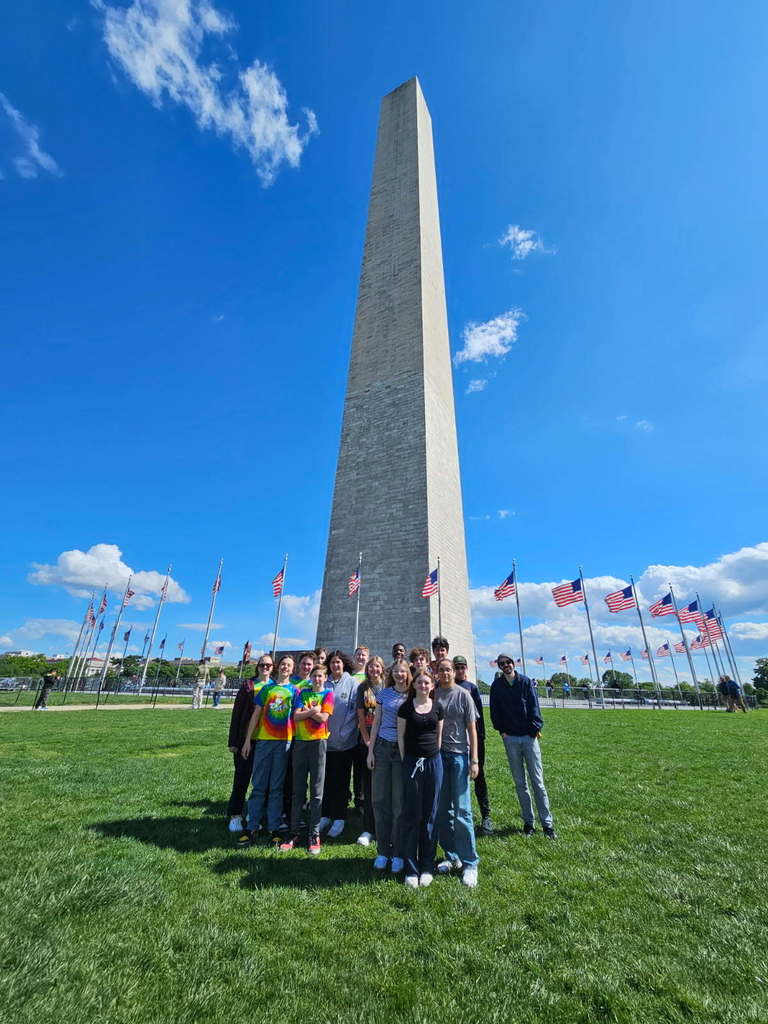 Junior High students and teachers stand together outdoors in front of the Washington Monument on a clear April day, smiling and posing for a group photo under a bright blue sky.