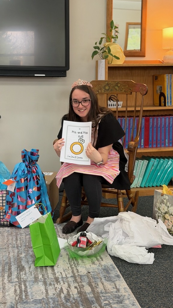 A smiling young woman sits in a wooden chair in a classroom, wearing a tiara and a striped apron over dark clothing. She holds up a handmade card that reads “Mr. and Mrs. Holt,” featuring a drawing of a ring. Around her on the floor are opened gift bags, tissue paper, and small presents, suggesting a celebration such as a bridal shower. Behind her are shelves filled with neatly arranged binders and books, a potted plant, and a softly lit lamp, creating a warm, welcoming environment.