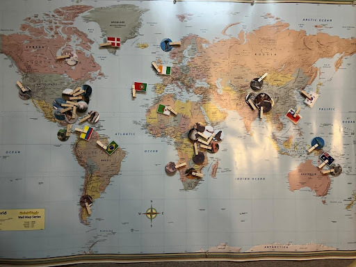 A large world map mounted on a classroom wall, with various small objects and flags pinned across different countries. The pins appear to represent locations or cultural connections, and some include miniature items or images attached to them. Sunlight casts soft shadows across the map, highlighting regions in North America, South America, Europe, Africa, Asia, and Australia.