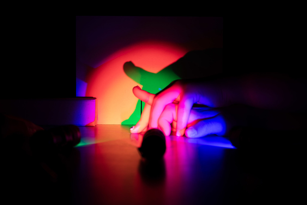 Close-up of a child’s hand illuminated by vivid red, blue, and green light while experimenting with colored flashlights and shadows against a glowing wall.
