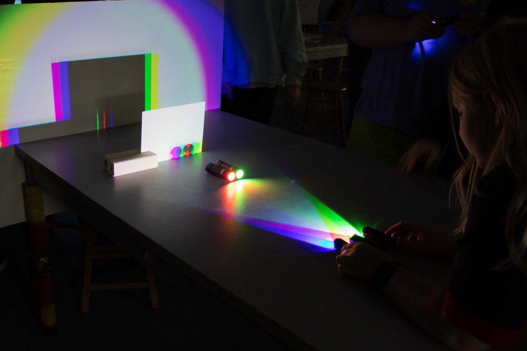 Children experiment with colored light and shadows in a darkened classroom, using flashlights and transparent materials to project vibrant rainbow patterns across a wall and table.