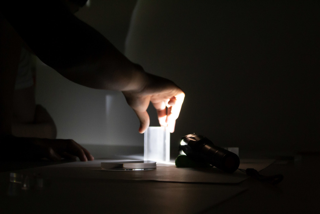 A student carefully positions a clear prism in front of a flashlight beam during a low-light science investigation exploring reflection and refraction.