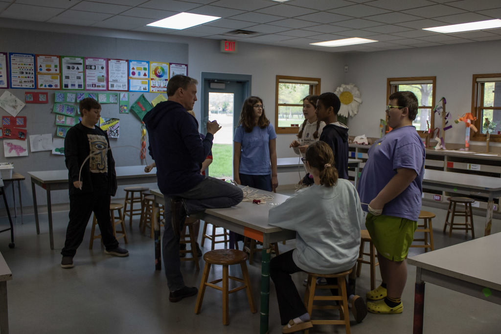 A teacher sits on a classroom table speaking with a group of students gathered around during a collaborative lesson in an art and science classroom filled with student artwork.