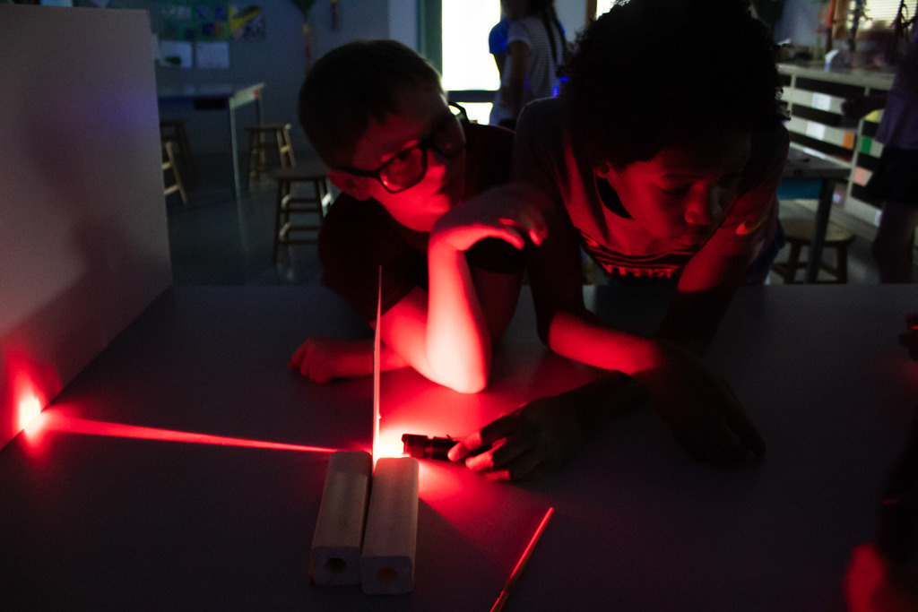Two students lean over a table in a darkened classroom while experimenting with a bright red laser beam and translucent materials during a light study activity.