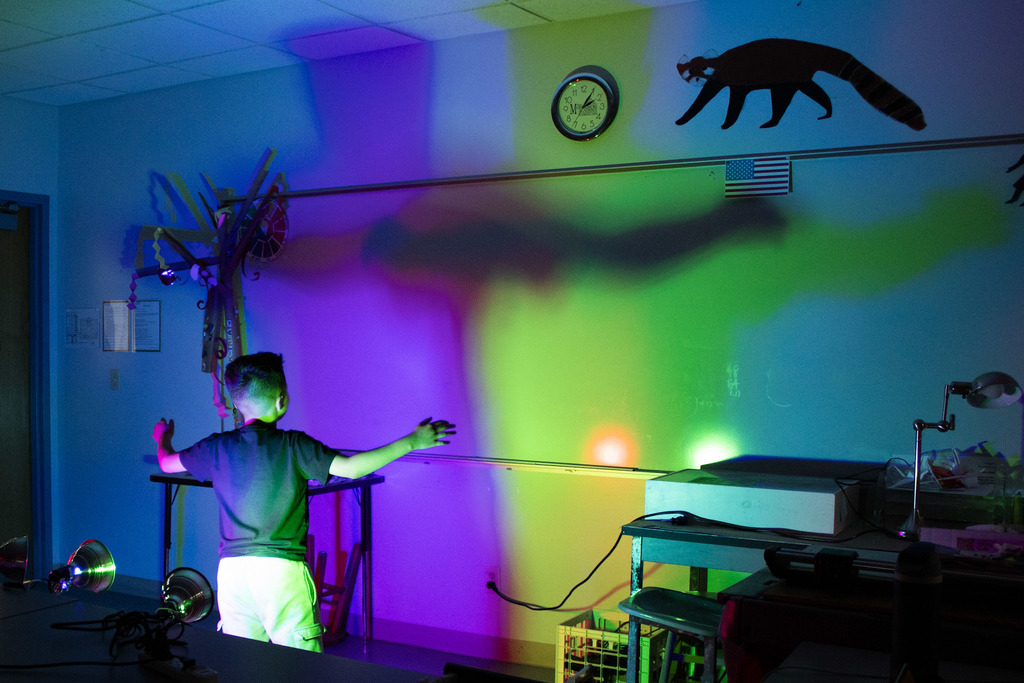 A young student stands with arms outstretched in front of a wall covered in overlapping rainbow-colored light projections and large shadows created during a classroom exploration of light and color.