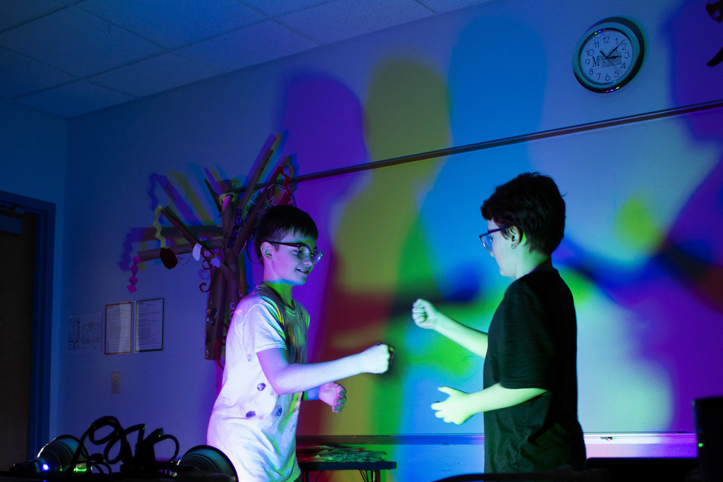 Two students stand facing one another in a dark classroom filled with colorful projected lights and shadows, smiling as they interact during a hands-on exploration activity.