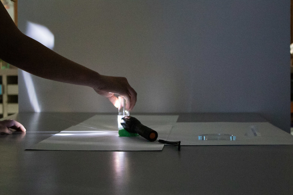 A student holds a clear prism in the beam of a flashlight during a hands-on light exploration activity, creating reflections and refractions across a tabletop in a dim classroom.