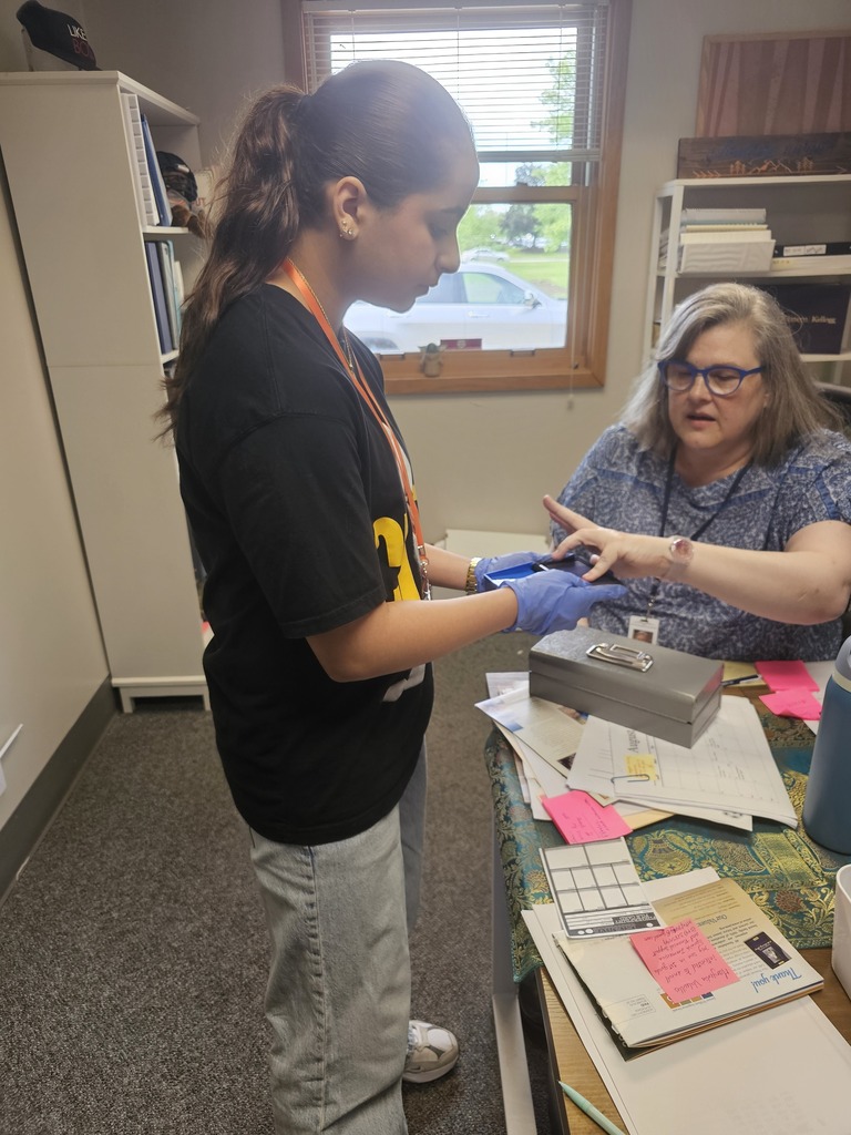 A Junior High student wearing gloves carefully examines and documents evidence while meeting with a staff member during a hands-on forensic science activity. Papers, notes, and investigation materials are spread across the desk.