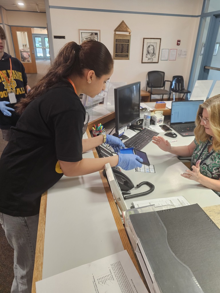 A Junior High student wearing gloves presents collected evidence to a staff member at the school reception desk during a forensic science investigation. Another student stands nearby observing the interaction.