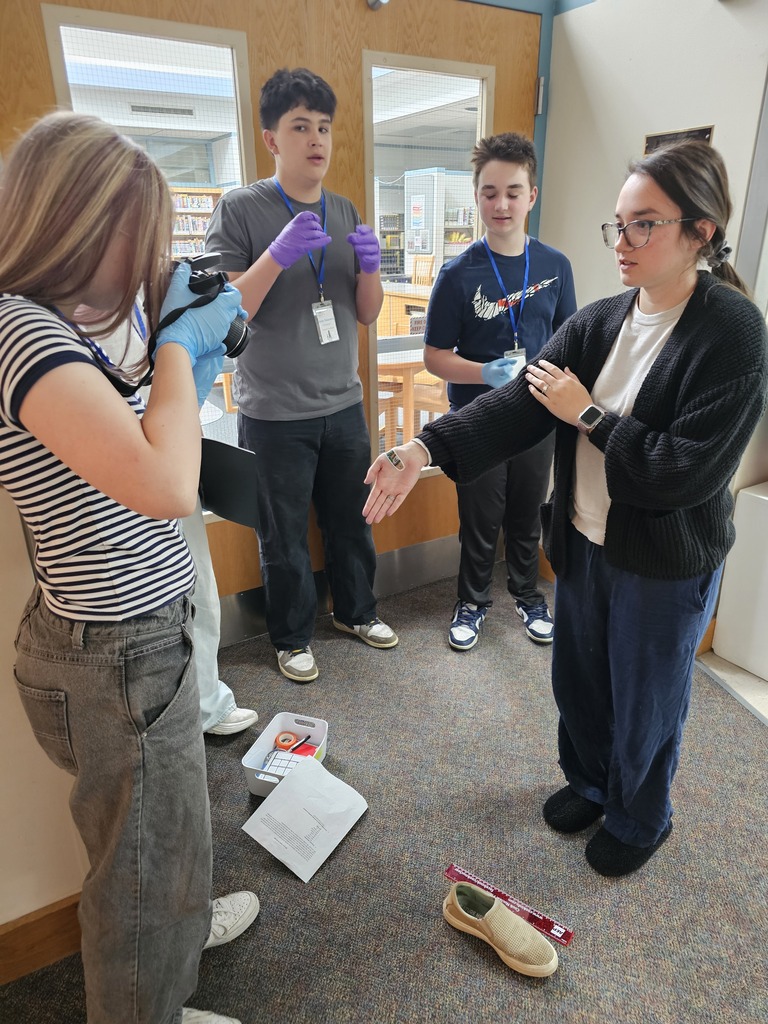 Junior High students wearing gloves and lanyards work together to document evidence during a forensic science investigation in the school hallway. One student photographs evidence markers while another student explains observations to the group.