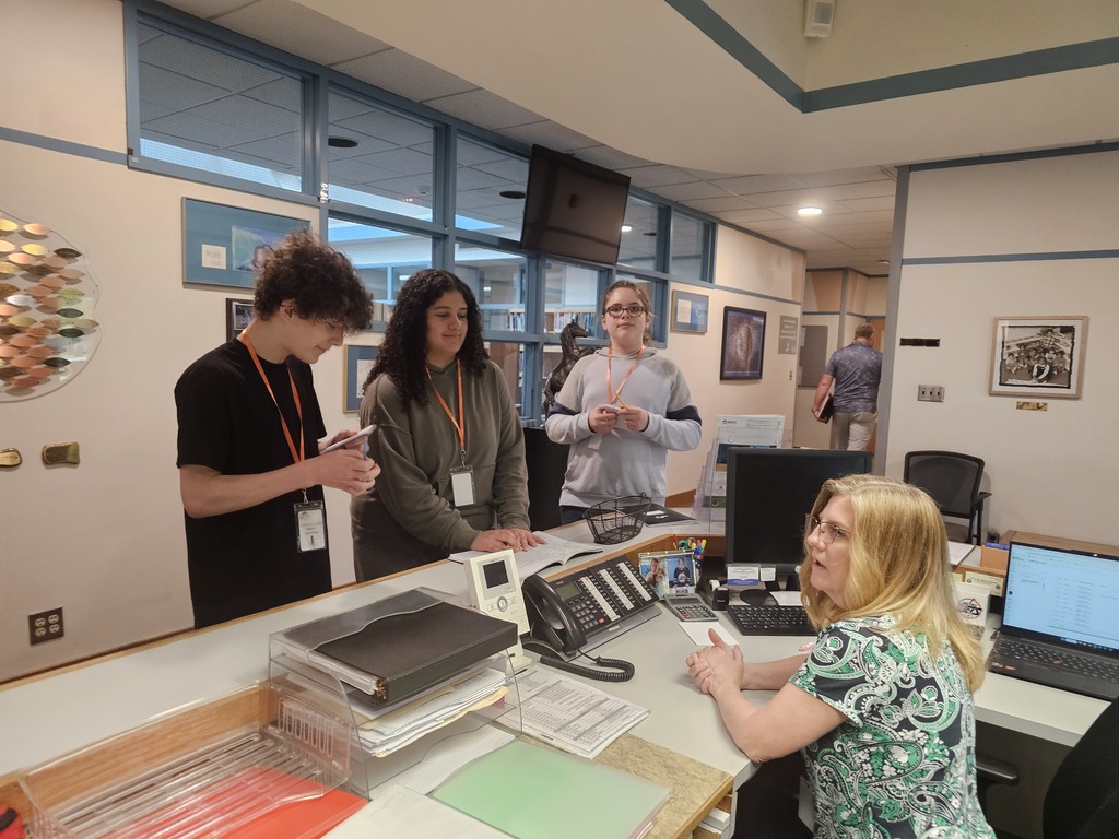 Junior High students interview a staff member at the front desk as part of their forensic science investigation. Students take notes and gather information while practicing observation and questioning skills.
