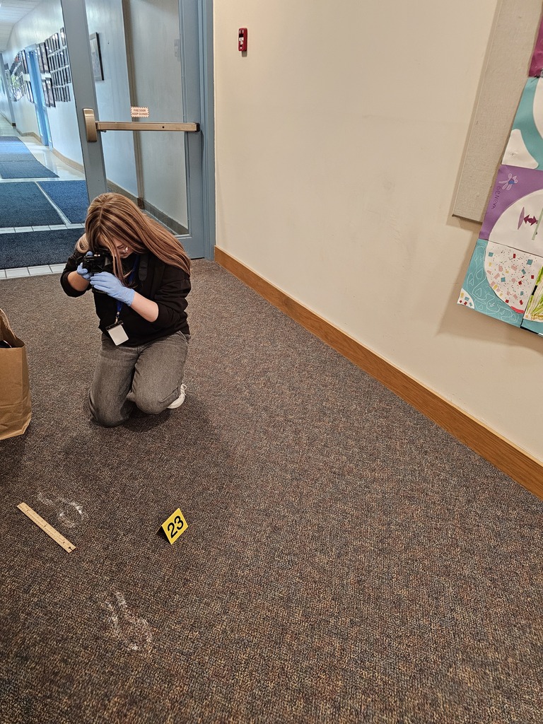 A Junior High student kneels in the hallway photographing evidence markers and footprints during a forensic science investigation. Measuring tools and evidence markers are placed carefully around the scene.