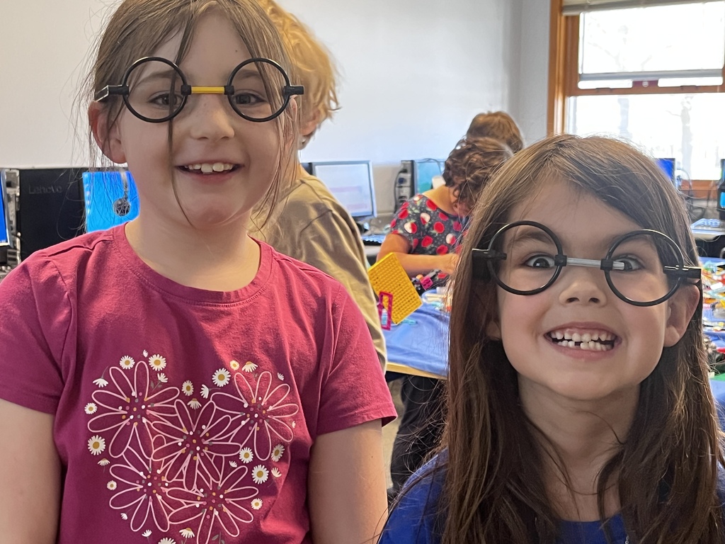 Two young students smile and pose playfully in a classroom while wearing oversized round glasses. One student wears a bright pink floral shirt, and the other wears a blue shirt with an excited expression. Other students and classroom materials can be seen working in the background.