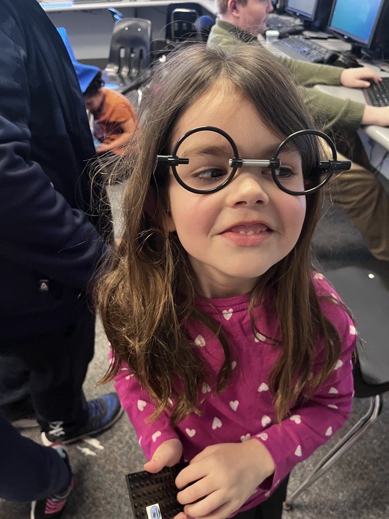 A young student wearing oversized round glasses smiles while standing in a classroom filled with students, computers, and activity tables. She holds a small black building piece while classmates work behind her.