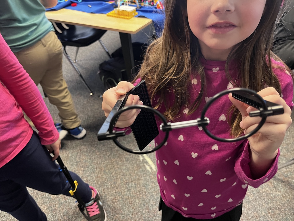 A young student in a pink heart-patterned shirt holds up a pair of oversized round glasses for the camera during a classroom activity. Other students and classroom tables are visible in the background.