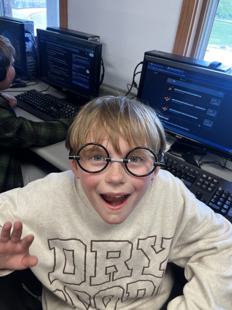 A smiling student wearing novelty round glasses poses excitedly in a computer lab classroom. Computer monitors and keyboards line the tables behind him while classmates work at nearby stations.