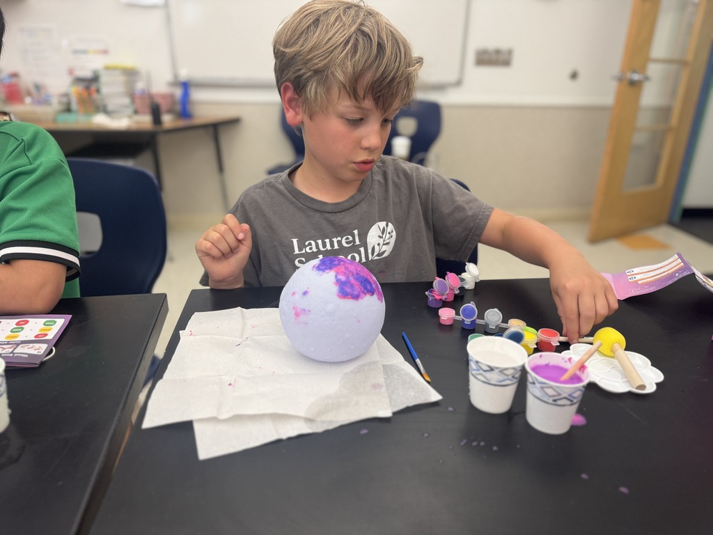 A child works on a craft project at a desk. The project involves a sphere with paint on it.