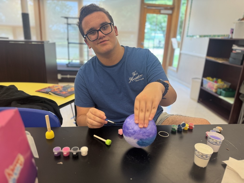 A person in a blue shirt sits at a desk, painting a spherical object. Behind them is a bookshelf.