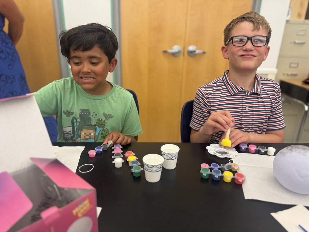 Two kids seated at a table with paint cups, a balloon, and various paint bottles. One is wearing a green shirt, the other a striped shirt.
