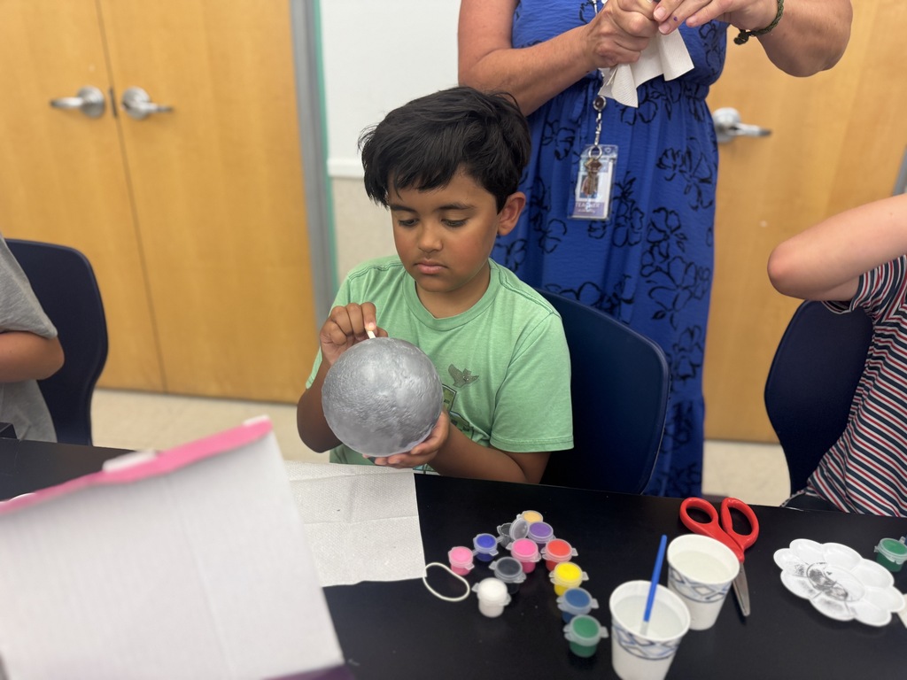 A young boy in green holds a silver ball, creating a craft. He is surrounded by a teacher, other students, and craft supplies.