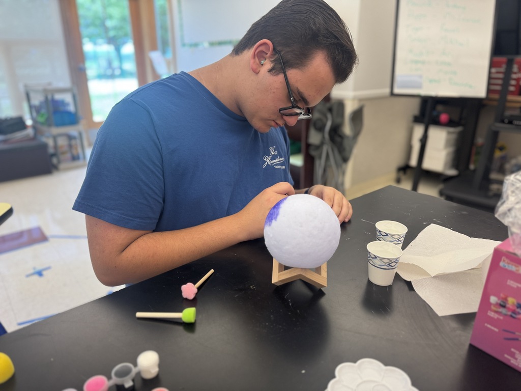 Person seated at a table, working on a painted sphere using a paintbrush and various colors.