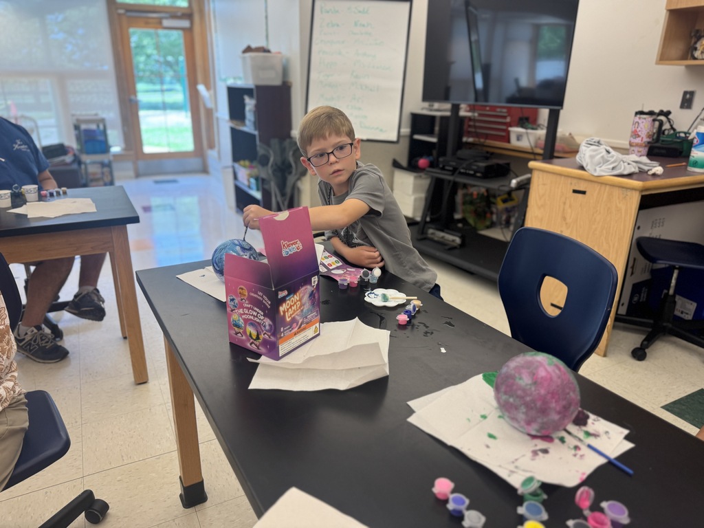 A boy in glasses works at a table in a classroom with a sphere, paint, and papers.