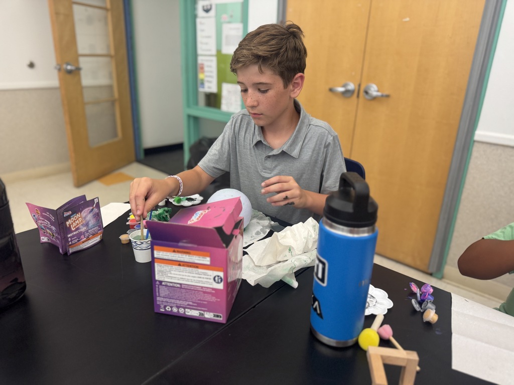 A boy in a grey shirt at a table with a purple box, a blue water bottle, and various objects.