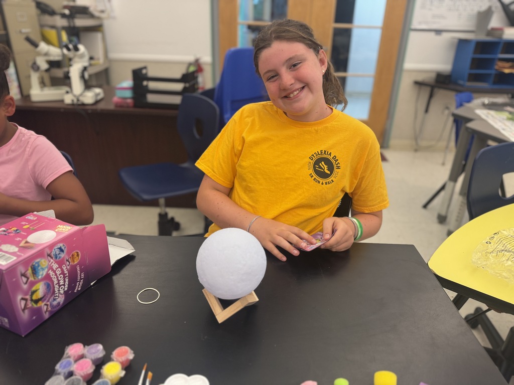 A young girl in a yellow shirt sits at a table, holding a white object. A box of art supplies and various colored paints are on the table.