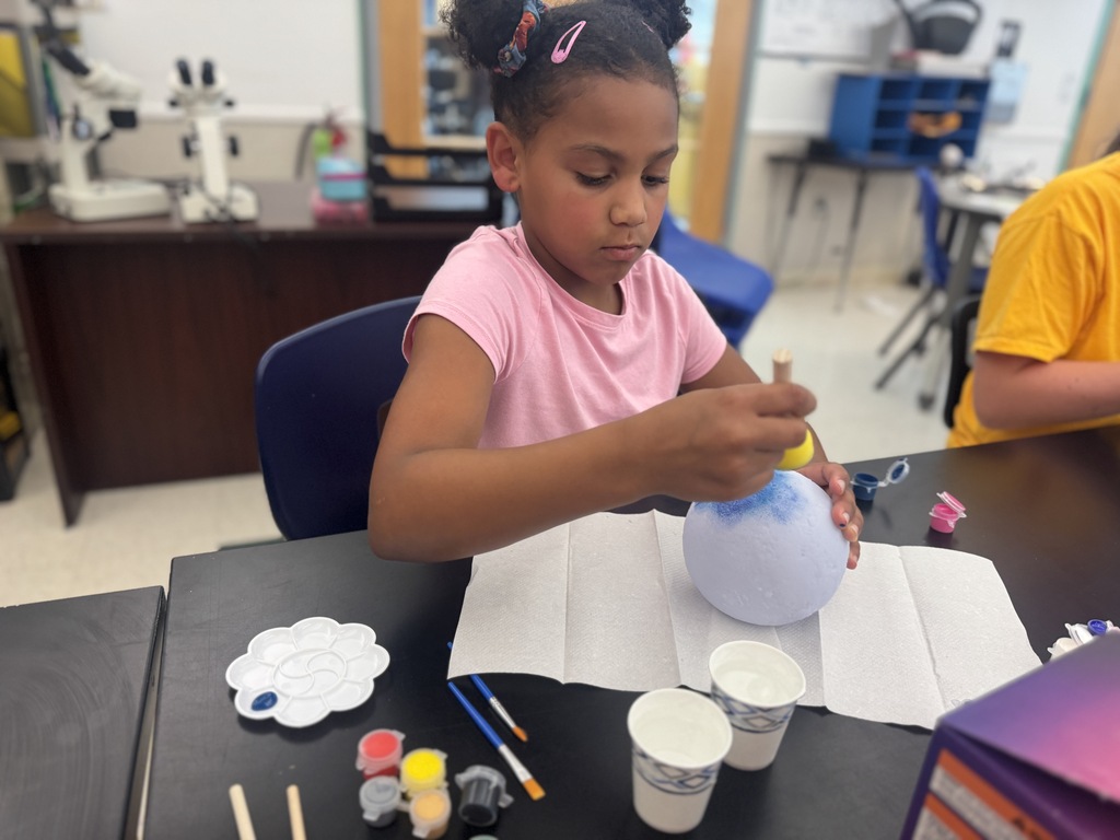 A young girl in a pink shirt uses a paintbrush to paint a white balloon at a table.