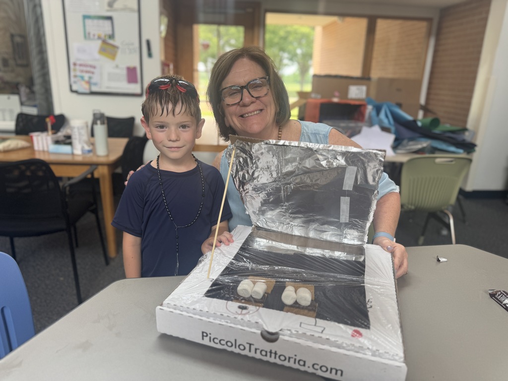 An adult and a child hold a box with a solar oven inside, standing at a table.
