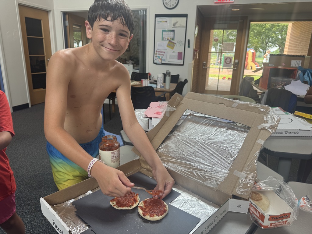 A young boy is opening a pizza box on a table in a room. He is holding a pizza and a pizza cutter.