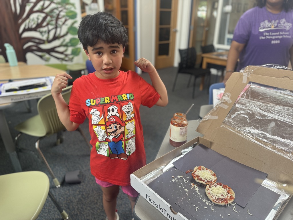 A young child in a red shirt stands next to a pizza box with pizzas inside.