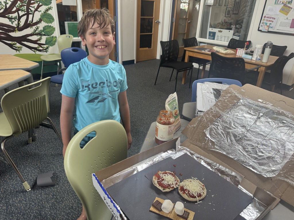 A boy stands in a classroom, next to a pizza in an open box. He wears a blue shirt.