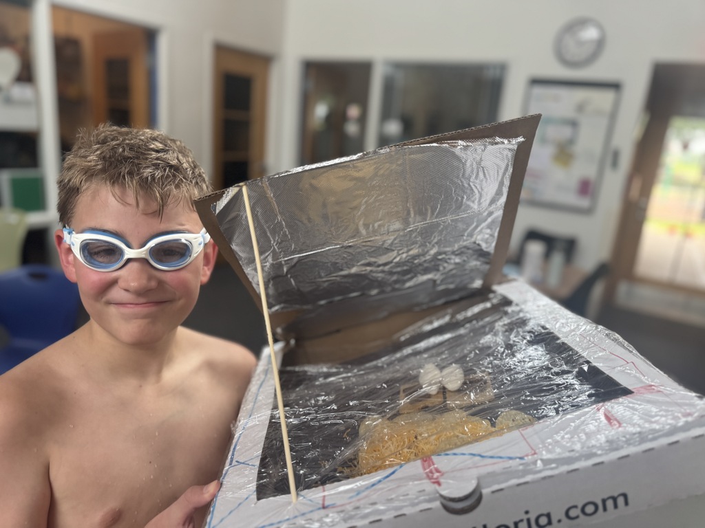 A young boy wearing goggles holds a homemade solar oven. The oven has foil on top.